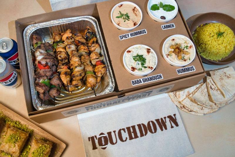 The Feast in a Football Feast box: a tray of chicken and beef kebobs beside four labeled dips, rice pilaf, pita, baklava, and cans of soda, on a Touchdown cloth