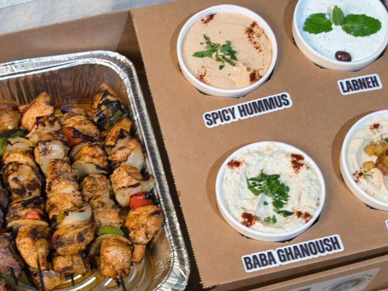 The Feast box: a tray of char-grilled chicken kebobs beside labeled cups of spicy hummus, labneh, baba ghannouj, and hummus